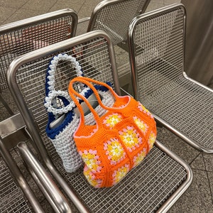 Op de afbeelding: Twee gebreide tassen, een witte met blauwe bies en een oranje met roze en gele accenten, hangen aan de rugleuning van een metalen stoel.