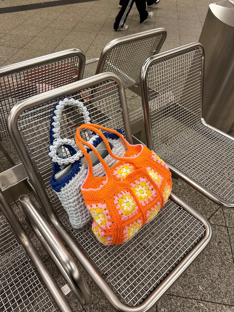 Op de afbeelding: Twee gebreide tassen, een witte met blauwe bies en een oranje met roze en gele accenten, hangen aan de rugleuning van een metalen stoel.