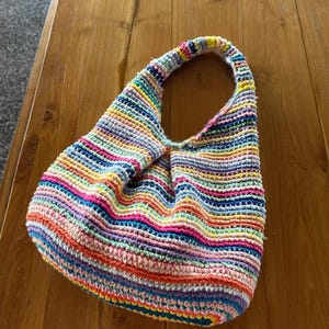 May include: A vibrant, hand-crocheted bag with a single handle. The bag displays horizontal stripes in a spectrum of bright colours, including pink, blue, yellow, and orange. The bag is resting on a wooden surface.