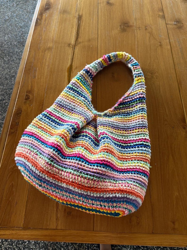 Puede incluir: Un bolso de ganchillo hecho a mano y colorido con un solo asa. El bolso presenta rayas horizontales en una variedad de colores brillantes, incluyendo rosa, azul, amarillo y naranja. El bolso est&aacute; sobre una superficie de madera.