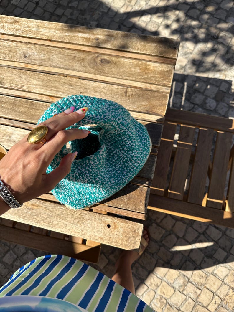 Pu&ograve; includere: Una borsa in crochet verde acqua e bianca con un motivo testurizzato. La borsa &egrave; tenuta da una mano che indossa un anello d'oro.