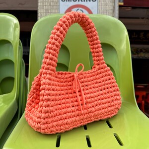 Pu&ograve; includere: Una borsa a mano all'uncinetto color corallo con manico arrotondato e chiusura con coulisse. La borsa &egrave; appoggiata su una sedia verde brillante. L'immagine mostra anche un cartello "NO SMOKING".