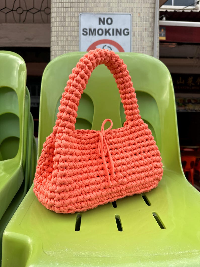 Pu&ograve; includere: Una borsa a mano all'uncinetto color corallo con manico arrotondato e chiusura con coulisse. La borsa &egrave; appoggiata su una sedia verde brillante. L'immagine mostra anche un cartello "NO SMOKING".