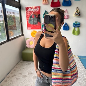 Puede incluir: Un bolso de hombro de ganchillo a rayas de colores. El bolso presenta una vibrante gama de colores del arco&iacute;ris. La imagen tambi&eacute;n muestra una colecci&oacute;n de peque&ntilde;os bolsos hechos a mano en varios colores y estilos exhibidos en una pared blanca.