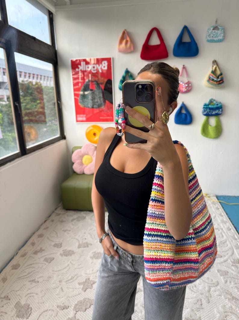 Puede incluir: Un bolso de hombro de ganchillo a rayas de colores. El bolso presenta una vibrante gama de colores del arco&iacute;ris. La imagen tambi&eacute;n muestra una colecci&oacute;n de peque&ntilde;os bolsos hechos a mano en varios colores y estilos exhibidos en una pared blanca.