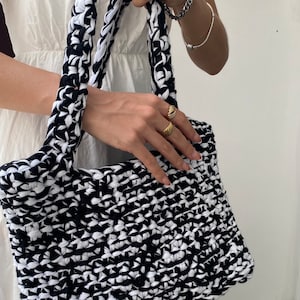 May include: A black and white crocheted tote bag with thick, chunky straps. The bag is made from recycled fabric and has a unique, textured design.