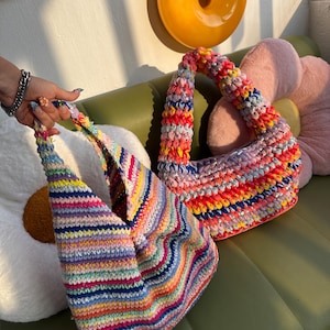K&ouml;nnte beinhalten: Zwei handgefertigte H&auml;keltaschen in leuchtenden Regenbogenfarben. Die Taschen haben ein gestreiftes Muster mit einer Vielzahl von Farben, darunter Rosa, Gelb, Blau und Gr&uuml;n. Die Taschen liegen auf einer gr&uuml;nen Oberfl&auml;che mit dekorativen Kissen.