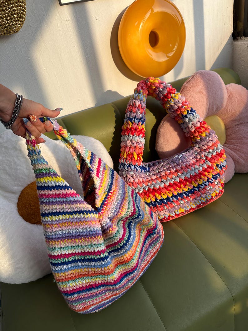K&ouml;nnte beinhalten: Zwei handgefertigte H&auml;keltaschen in leuchtenden Regenbogenfarben. Die Taschen haben ein gestreiftes Muster mit einer Vielzahl von Farben, darunter Rosa, Gelb, Blau und Gr&uuml;n. Die Taschen liegen auf einer gr&uuml;nen Oberfl&auml;che mit dekorativen Kissen.