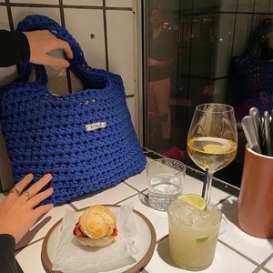 May include: A blue crocheted tote bag with the word "ByGALK" on the front. The bag is sitting on a table with a glass of white wine, a glass of a mixed drink, and a sandwich on a plate.