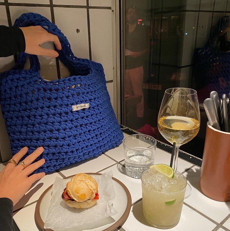 May include: A blue crocheted tote bag with the word "ByGALK" on the front. The bag is sitting on a table with a glass of white wine, a glass of a mixed drink, and a sandwich on a plate.