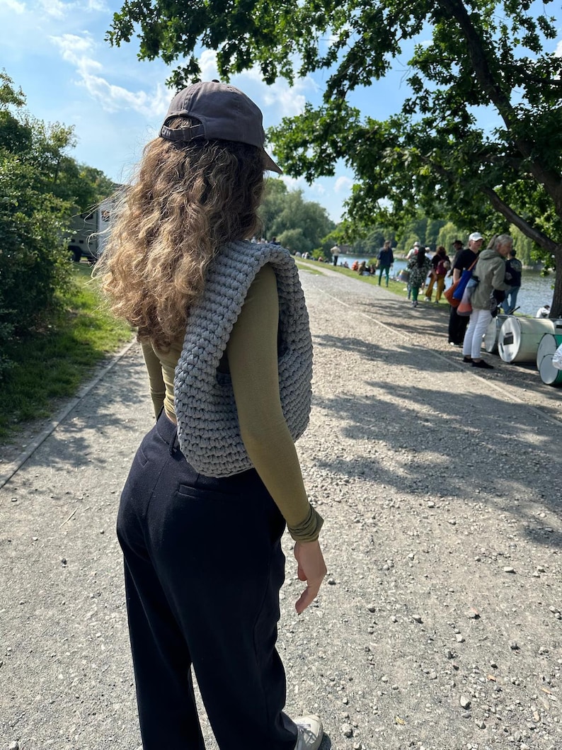 May include: A person wearing a gray baseball cap, a green long-sleeved shirt, and black pants. They are carrying a gray crocheted bag.
