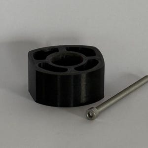 Op de afbeelding: Een zwart 3D-geprint object van plastic met een zeshoekig gat in het midden en vijf kleinere gaten rondom de omtrek. Een zilveren metalen schroef ligt naast het object.