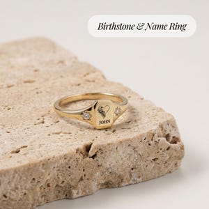 Op de afbeelding: Een goudkleurige ring met een rechthoekig vlak gegraveerd met de naam JOHN en een hartsymbool. De ring is aan weerszijden van de centrale plaat versierd met twee kleine, heldere, stervormig gezette steentjes voor een gepolijste afwerking.