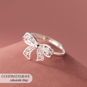 Op de afbeelding: Een zilveren verstelbare ring met een strik ontwerp. De strik heeft ingewikkeld opengewerkt en een klein, rond midden. De ring wordt getoond tegen een roze achtergrond. Tekst op de afbeelding luidt "CUSTOM ENGRAVE Adjustable Ring."