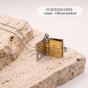 Op de afbeelding: Een roestvrijstalen boekvormige medaillonketting met een zilverkleurige ketting. Het medaillon opent en toont goudkleurige pagina's met de gegraveerde tekst: She believed she could, so she did. Het medaillon heeft een kleine hartvormige uitsparing.