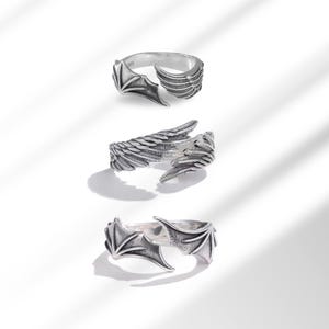 May include: Three silver rings with wing designs. The top ring features a bat wing design on one side and a feathered wing on the other. The middle ring has a feathered wing design. The bottom ring has a bat wing design.