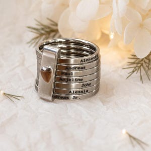 May include: A stack of silver-toned metal rings held together by a vertical bar featuring a small heart emblem. Each individual ring is engraved with a name, including Alexandre, Andreas, Paul, Anjeline, John, Alessia, and Andreas Jr.