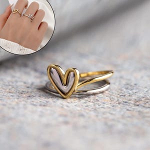 May include: A gold and silver heart-shaped ring. The ring features a gold heart outline with a silver interior, set on a double band. A smaller inset image shows two other rings, one gold heart-shaped and one silver with a V-shape.
