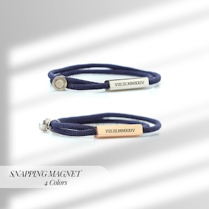 Op de afbeelding: Twee marineblauwe touwarmbanden met rechthoekige metalen sluitingen. De ene sluiting is zilverkleurig, de andere goudkleurig. Elke sluiting heeft Romeinse cijfers gegraveerd. De tekst "SNAPPING MAGNET" en "4 Colors" staat onderaan.
