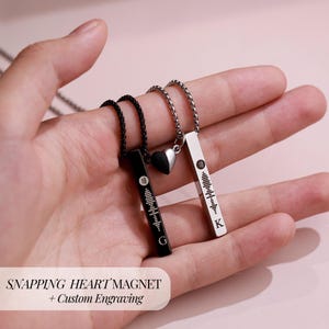 May include: Two matching necklaces with bar pendants and a heart-shaped magnet. One pendant is black with the letter "G", the other is silver with the letter "K". Each pendant has a unique sound wave design. The text "SNAPPING HEART MAGNET + Custom Engraving" is at the bottom.