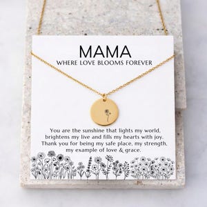 Könnte beinhalten: Eine goldfarbene Halskette mit einem runden Anhänger mit floralem Design. Der Anhänger hängt an einer zarten Kette. Die Halskette wird auf einer weißen Karte mit dem Wort "MAMA" und dem Satz "WHERE LOVE BLOOMS FOREVER" präsentiert.