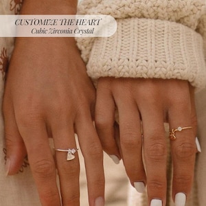 May include: Two hands wearing delicate rings. One ring is silver with a heart charm and a cubic zirconia crystal. The other ring is gold with a small heart charm and a single stone. The text in the image reads "CUSTOMIZE THE HEART Cubic Zirconia Crystal."