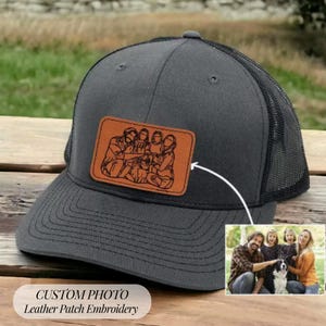 Puede incluir: Una gorra tipo trucker de color gris carbón con un parche rectangular de cuero color canela en la parte frontal. El parche muestra una ilustración de líneas marrón oscuro de una familia de cuatro personas con un perro, basada en la foto.