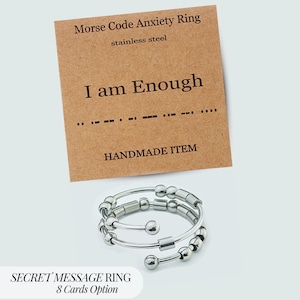 May include: A stainless steel anxiety ring with a Morse code design, accompanied by a card that reads "I am Enough." The ring features a spiral design with small, movable beads. The card is brown with black text. The product is a handmade item.