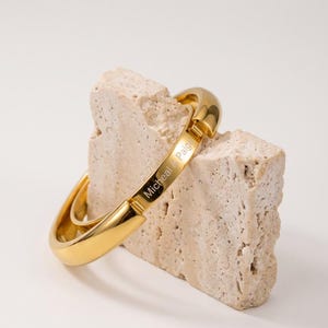 May include: A polished gold-tone metal hinged bangle bracelet with a smooth, rounded surface. The bracelet features an engraved rectangular plaque on the front with the text Micheal & Paige clearly visible in a clean, sans-serif font.