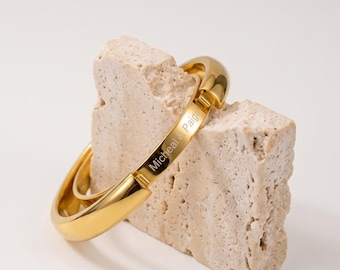 May include: A polished gold-tone hinged bangle bracelet featuring a smooth, rounded surface. The metal band is engraved with the text Micheal & Paige on the flat inner section, showcasing a clean, reflective finish and a secure clasp mechanism.
