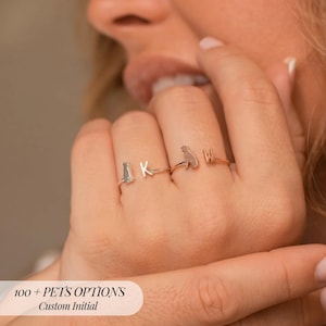 Puede incluir: Cuatro anillos de plata con diseños temáticos de mascotas e iniciales personalizadas. Los anillos presentan un gato, la letra "K", un perro y la letra "W". El texto "100 + PETS OPTIONS Custom Initial" es visible en la parte inferior de la imagen.