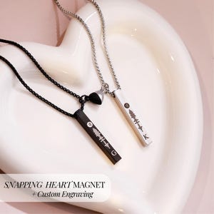 May include: Two silver necklaces with a heart charm and a bar pendant engraved with a music note and a custom message. The necklaces are on a white heart-shaped surface. "SNAPPING HEART' MAGNET + Custom Engraving" is written below the necklaces.