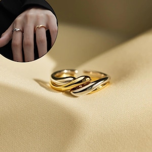 May include: A gold and silver ring with a distinctive, fluid design. The ring has a layered, open-ended style with smooth, curved bands. The close-up shot highlights the polished finish and elegant aesthetic of the ring.