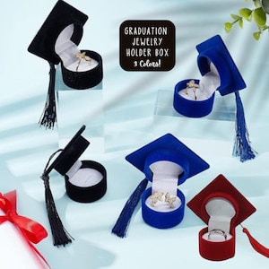Può includere: Tre scatole per anelli a forma di cappello di laurea in velluto nero, blu e rosso. Le scatole sono aperte e mostrano un anello, orecchini e un paio di orecchini all'interno. Il testo "GRADUATION JEWELRY HOLDER BOX 3 Colors!" è visibile nell'immagine.