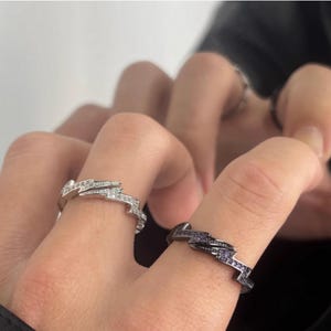 Pode incluir: Dois anéis com um design de raio. Um anel é prateado com pedras transparentes, e o outro é preto com pedras roxas. Os anéis estão nos dedos de uma pessoa.