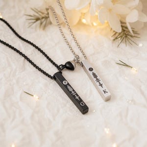 May include: Two bar pendant necklaces on chains, one black and one silver. Each bar features a laser-engraved sound wave pattern, a small circular icon, and a single letter at the bottom, with the letter G on the black bar and K on the silver bar.