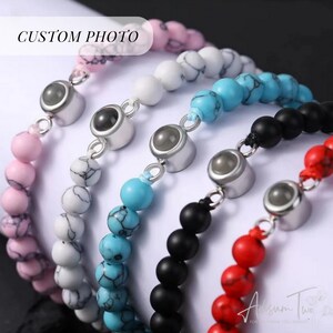 May include: Five beaded bracelets with a silver charm featuring a small round lens. The bracelets are pink, white, turquoise, black, and red.