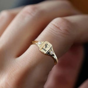 May include: A gold-colored ring with a rectangular face featuring the name "JOHN" and a heart-shaped design with a horse. The ring has two small, clear gemstones on either side of the face.