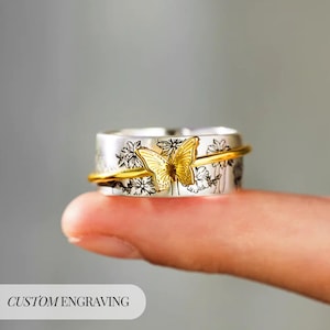 May include: A silver ring with a gold butterfly and a gold band. The ring features a floral design and the text "CUSTOM ENGRAVING". The butterfly is centered on the ring, with its wings spread.