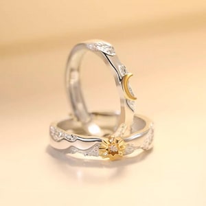May include: Two silver rings with celestial designs. One ring features a gold sun with a central stone, and the other has a gold crescent moon. Both rings have a wave-like pattern and small stones.