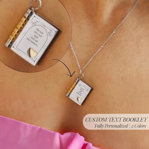 Peut inclure: Un collier argenté avec un pendentif en forme de livre. Le pendentif livre a des accents dorés et un détail en forme de cœur. Le texte sur le livre dit : "She was fire, and light and ash and embers...". Le collier est présenté sur un fond de peau.