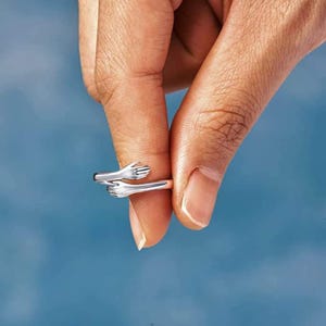 May include: A silver-colored ring with a design of two hands embracing. The ring is shown against a blue background. The hands are detailed with fingers and are wrapped around each other.