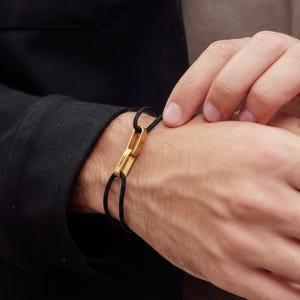 May include: A black cord bracelet with a gold-colored rectangular clasp. The clasp has the engraved text "DIMMEL" and "LIA". The bracelet is worn on a wrist, with a black jacket in the background.