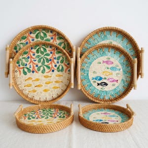 May include: A collection of decorative rattan trays with colorful mosaic designs. The trays feature painted fish and floral patterns in various sizes, with wooden handles for easy carrying. The color palette includes blue, green, yellow, and beige.