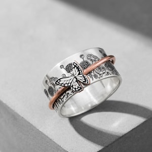 May include: A silver ring with a butterfly design. The ring features a copper-colored band and is decorated with a black and white butterfly and floral engravings. The ring is a fashion accessory.