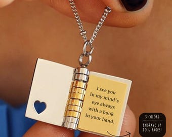 Può includere: Collana color argento con un ciondolo a forma di libro in miniatura. Il libro ha un ritaglio a forma di cuore e una pagina gialla con il testo "I see you in my mind's eye always with a book in your hand." Il ciondolo è tenuto da una persona.
