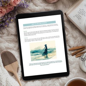 Op de afbeelding: Een digitale tablet met een journaalpagina met instructies en een aquarel illustratie van een persoon op een strand. De afbeelding is omgeven door bloemen, een kaars, een boek en een kopje thee.