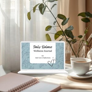 Op de afbeelding: Een laptop toont een "Daily Balance Wellness Journal" met een bloemreli&euml;fpatroon. Een roze notitieboek en een witte theekop op een schotel staan op een houten oppervlak. Groene planten en een transparant gordijn zijn op de achtergrond.