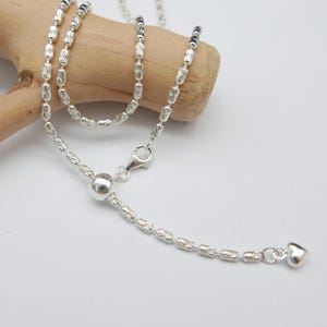 May include: A delicate silver necklace with a small heart pendant and a ball-shaped accent. The necklace is displayed on a light-coloured wooden branch against a white background. The necklace features a lobster clasp.