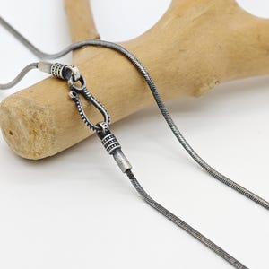 May include: A silver-coloured necklace with a snake chain design. The necklace features an ornate clasp and a decorative pendant. The jewellery is displayed against a light background with a wooden element.